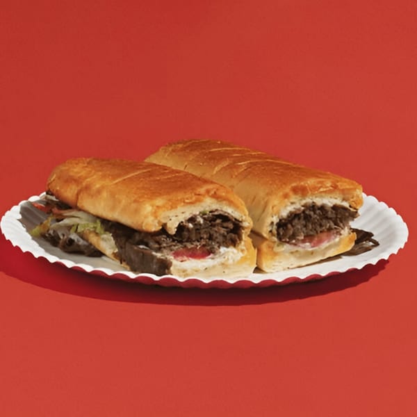 Two steak and cheese subs on a white paper plate against a red background.