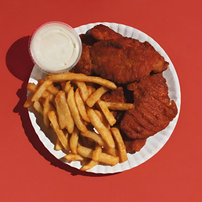 A white paper plate holds crispy french fries, red chicken tenders, and a small cup of dipping sauce.