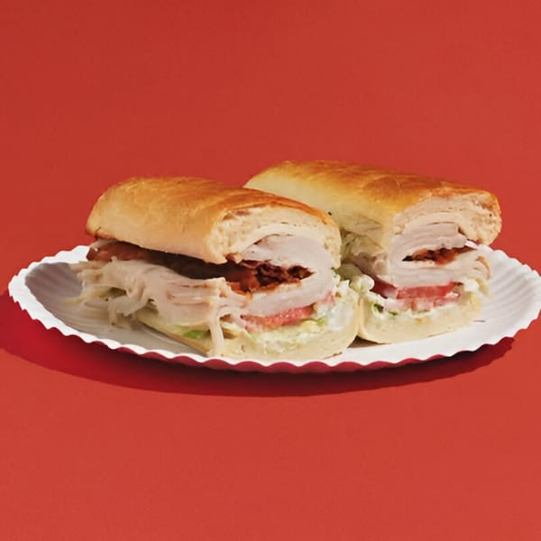 Two turkey and bacon subs with lettuce, tomato, and mayo on a white paper plate.