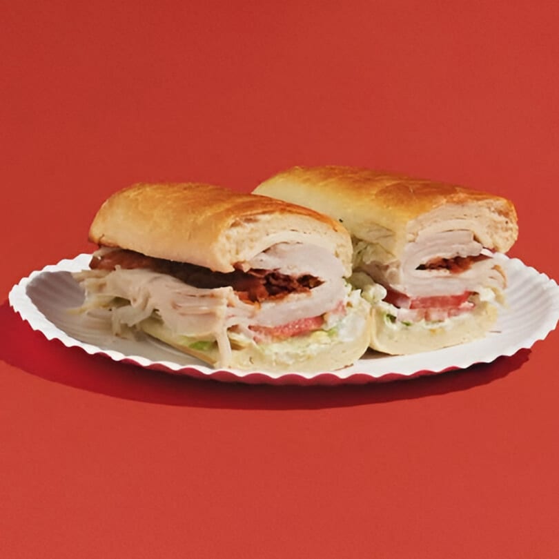Two turkey and bacon subs with lettuce, tomato, and mayo on a white paper plate.
