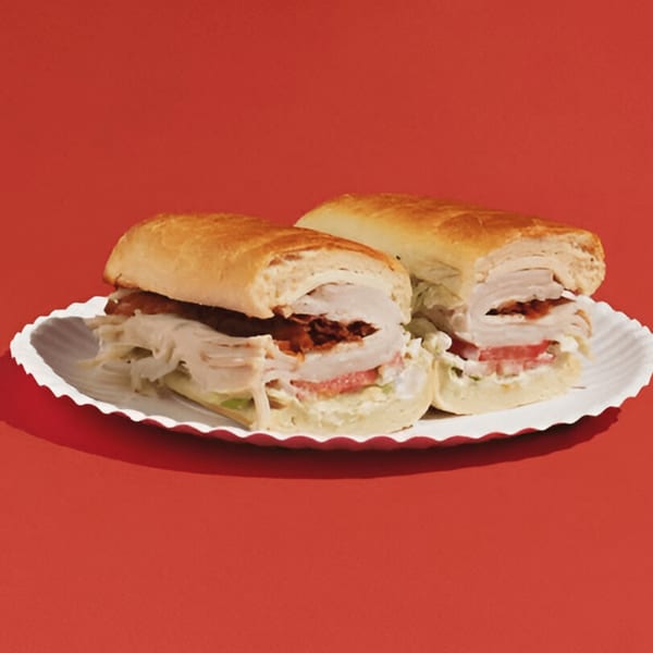Two turkey and bacon subs with lettuce, tomato, and mayonnaise on a white paper plate.