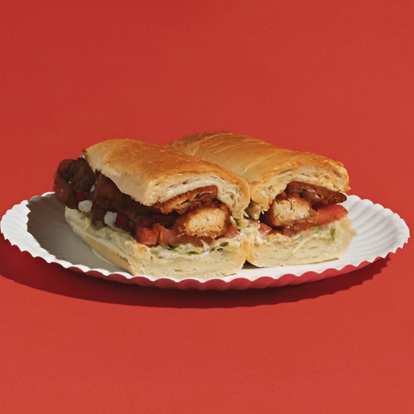 Two chicken sandwiches with lettuce and tomato on a white paper plate against a red background.